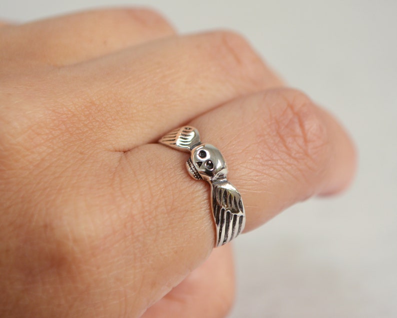 May include: A silver ring featuring a skull with detailed wings. The ring is worn on a finger, showcasing the intricate design and craftsmanship. The skull has detailed eye sockets and a small nose.