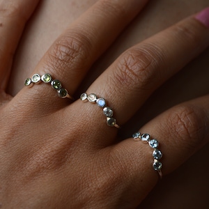 May include: Three silver rings with different coloured gemstones. The top ring has green gemstones, the middle ring has white gemstones, and the bottom ring has blue gemstones.