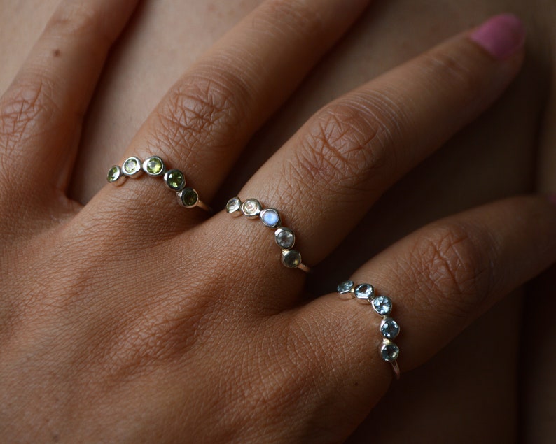 May include: Three silver rings with different coloured gemstones. The top ring has green gemstones, the middle ring has white gemstones, and the bottom ring has blue gemstones.