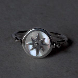 May include: A silver ring with a mother-of-pearl face, displaying a starburst design. The ring has a polished finish and a simple band with small ball accents. The ring is set against a dark grey background, with a focus on the ring's details.