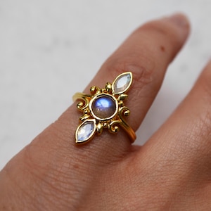 May include: A gold ring featuring a large round moonstone and two smaller moonstones on either side. The ring has a delicate, ornate design.