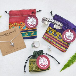 May include: Four small drawstring bags made of red, purple, green, and dark green fabric. Each bag has a round tag with the words "mangosteen dreams" and a small image of a mango. The bags contain a silver elephant necklace, a silver bracelet with the text "ओम नमः शिवाय" in Hindi, a turquoise ring, and a silver ring.