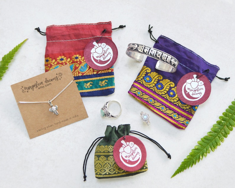 May include: Four small drawstring bags made of red, purple, green, and dark green fabric. Each bag has a round tag with the words "mangosteen dreams" and a small image of a mango. The bags contain a silver elephant necklace, a silver bracelet with the text "ओम नमः शिवाय" in Hindi, a turquoise ring, and a silver ring.