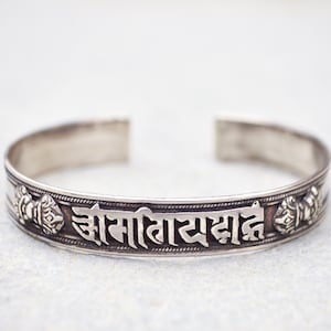 Puede incluir: Un brazalete plateado con texto grabado y elementos decorativos. El brazalete abierto presenta patrones e inscripciones detalladas, mostrando un diseño tradicional. El brazalete se exhibe sobre una superficie clara.