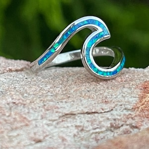 May include: A sterling silver wave ring with a blue and green opal inlay. The ring's design mimics a curling ocean wave, with the opal adding a splash of color. The ring is set against a blurred green background.
