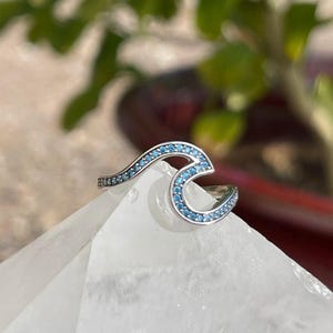 May include: A silver wave ring with blue gemstones. The ring is set on a white crystal surface.