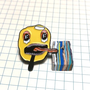 May include: Yellow enamel pin with a cartoon face with a large open mouth and a long red tongue sticking out. The face has two large black eyes with white pupils and red eyelids. The pin is on a white grid background.