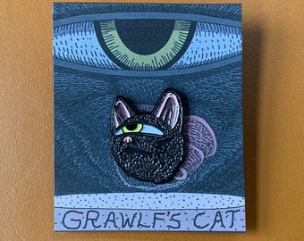 May include: A black enamel pin featuring a cartoon cat with a single large eye. The cat is depicted against a dark blue background with a large eye in the background. The text "GRAWLF'S CAT" is printed below the cat.
