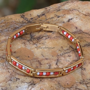 Puede incluir: Pulsera dorada con diseño de eslabones rectangulares. Los eslabones están adornados con cuentas rojas y blancas alternas. La pulsera se muestra sobre una superficie de piedra marrón claro texturizada. El texto "spellangelempoum" es visible.