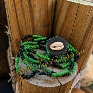 May include: A multi-strand beaded bracelet featuring alternating black and green glass beads. The bracelet includes a central circular black disc adorned with a single white cowrie shell. The text Spell Angel Emporium is visible.