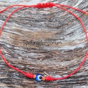May include: Red string bracelet with an evil eye charm. The bracelet features a small blue and white eye bead, with gold-colored beads on either side. The string is knotted at the ends for adjustability. The text "Spellangelemporium" is visible.