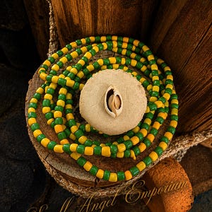 May include: A strand of alternating yellow and green beads coiled around a circular, light beige textured disc. A single small, white, oval-shaped cowrie shell is centered on the surface of the disc. The text Spell Angel Emporium is visible.