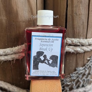 May include: A small, rectangular glass bottle filled with red liquid, labelled "Fragancia de Aceite Scented Oil Separacion Break Up." The label features a silhouette of two figures. The bottle has a white cap and is displayed against a wooden background.