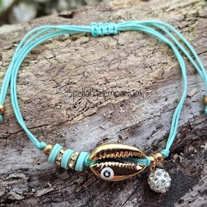 May include: A turquoise adjustable bracelet featuring a gold-colored seashell charm with an evil eye design, accented with gold beads and a sparkling ball charm. The bracelet also includes turquoise beads and is set against a natural wood background.