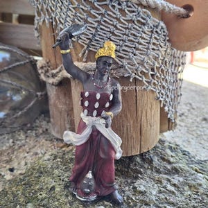 May include: A figurine of a person in a maroon dress with white polka dots and a gold crown. The figure is holding a blade in one hand and a small object in the other. The background includes a wooden post, fishing net, and a stone surface.