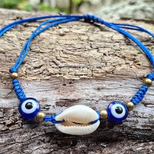 May include: A blue cord bracelet featuring a white seashell centerpiece, flanked by two blue and white evil eye beads. The bracelet includes gold-colored beads and is set against a textured wooden surface.