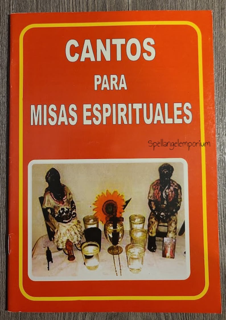 Pu&ograve; includere: Un libro rosso con il titolo "Cantos Para Misas Espirituales" e un bordo giallo. La copertina presenta una foto di un tavolo con due figurine, candele e un girasole.