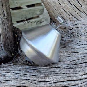 May include: A silver-colored, geometric bottle stopper with a small handle. The stopper has a twisted, angular design. The background is a weathered, gray wooden surface.