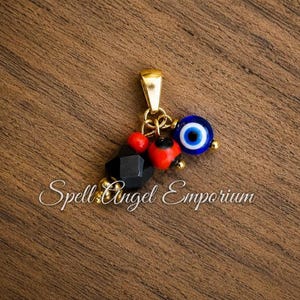 May include: A gold-toned metal pendant featuring a cluster of beads including a blue and white evil eye bead, a faceted black bead, and two small red beads, all suspended from a gold-toned bail. The text Spell Angel Emporium is visible.