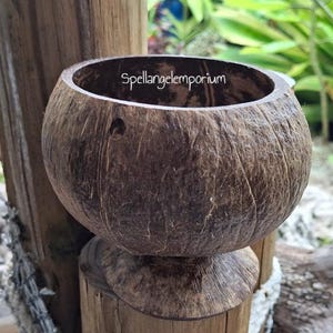 Puede incluir: Un cuenco artesanal de cáscara de coco marrón con forma redondeada y base de pedestal. El cuenco tiene una superficie natural y texturizada y el texto "Spellangelemporium" es visible en el borde. El cuenco se exhibe en un poste de madera.