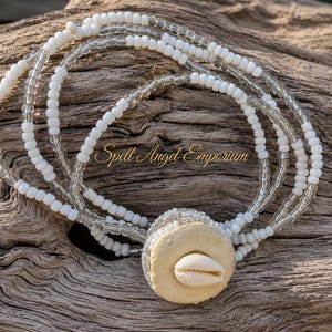 May include: A multi-strand bracelet featuring alternating white and clear glass beads. The bracelet includes a central circular beige disc adorned with a single white cowrie shell. The text Spell Angel Emporium is visible in the center of the image.