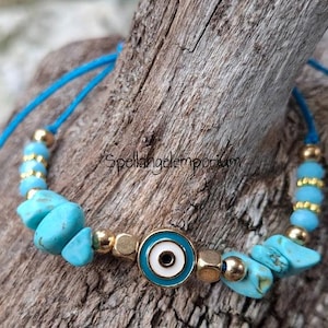 May include: A turquoise and gold beaded bracelet with an evil eye charm. The bracelet features turquoise stone chips, gold beads, and a blue cord. The evil eye charm is white and blue with a gold border. The text "Spellangelemporium" is visible.