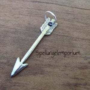 Puede incluir: Colgante de flecha plateada con un diseño detallado de plumas y una pequeña gema negra. La flecha está en ángulo diagonal sobre una superficie de madera. El texto "spellangelemporium" es visible.