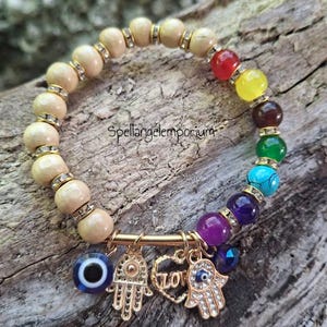 May include: A beaded bracelet with wooden beads and colorful gemstone beads. The bracelet features gold charms, including a Hamsa hand, an eye charm, and a heart with the word "LOVE". The bracelet is displayed on a wooden surface.