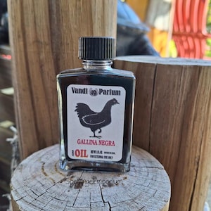 以下が含まれることがあります： 黒い鶏のイラストと「Gallina Negra」の文字が入った黒いラベルが付いた、小さな正方形のガラス瓶の「OIL」。ボトルには黒いキャップが付いており、木製の表面に置かれています。「Vandi Parfum」の文字も表示されています。