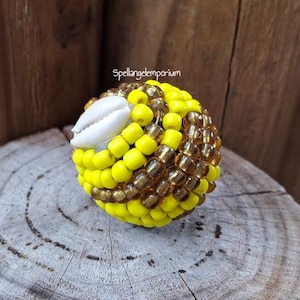 May include: A beaded sphere with yellow and brown beads, and a white seashell accent. The sphere is sitting on a wooden surface. The text "Spellangel Emporium" is visible in the background.