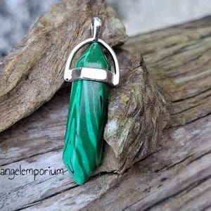May include: A green malachite crystal pendant with a silver bail. The pendant is on a piece of driftwood. The text "Spellangelemporium" is visible in the lower left corner of the image.
