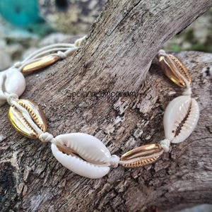 May include: A handmade bracelet featuring natural white seashells and gold-colored accents. The shells are strung together with a cream-colored cord. The bracelet is displayed on a piece of driftwood. The text "Spellangelemporium" is visible.