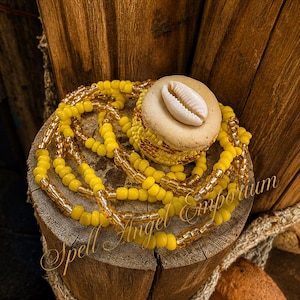 May include: A waist bead strand featuring bright yellow and translucent gold glass beads, coiled around a circular tan base topped with a single white cowrie shell. The text Spell Angel Emporium is overlaid in a cursive script across the image.