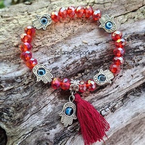 May include: Red beaded bracelet with silver Hamsa hand charms and a red tassel. The bracelet features faceted red beads and a dangling Hamsa charm with a blue eye. The bracelet is displayed on a wooden surface.
