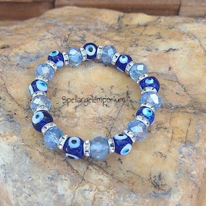 May include: A blue and white evil eye bracelet with crystal beads. The bracelet is on a light brown rock.