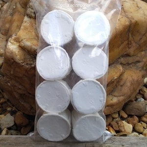 May include: A clear plastic bag filled with eight white, round, disc-shaped objects. The objects have a slightly textured surface and are stacked in two rows of four. The background includes rocks and wood.