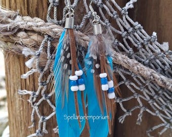 Op de afbeelding: Handgemaakte oorhangers met veren. De oorbellen hebben heldere turquoise veren, bruine leren accenten en zilverkleurige haken. Witte en blauwe kralen zijn geregen langs het leer. De tekst "Spellangelemporium" is zichtbaar op de veren.