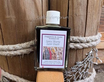 May include: A small, rectangular bottle of scented oil with a white cap. The label reads "Fragancia de Aceite Scented Oil Santa Muerte Saint Death." The bottle is set against a wooden background with rope detailing.