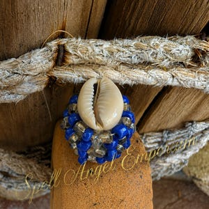 May include: A ring featuring a central white cowrie shell surrounded by a cluster of small, vibrant blue and clear translucent beads. The beadwork is arranged in a rounded, textured pattern, creating a circular band that encircles the shell.