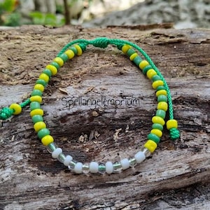 Puede incluir: Una pulsera de cuentas con cuentas verdes, amarillas y blancas. La pulsera está ensartada en un cordón verde y presenta el texto "Spellangelemporium". La pulsera se muestra sobre un trozo de madera.