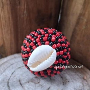 May include: A round, beaded object with red and black beads, and a white seashell. The object is resting on a textured surface. The text "Spellangelemporium" is visible on the bottom right.