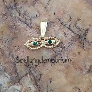 May include: Gold pendant with two green eyes, resembling a mask. The pendant has a rope-like design around the eyes.