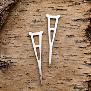May include: Two silver-toned metal charms shaped like medical crutches. Each charm features a tapered design with a horizontal bar and two small protrusions at the top, finished with a rounded tip at the bottom. The text SpellAngelEmporium is visible.