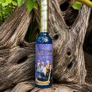 May include: A tall, cylindrical glass bottle filled with bright blue cologne. The label features the text Llama Cliente and a graphic of four people in an office setting. The bottle has a gold foil neck seal with the word ORIGINAL repeated.