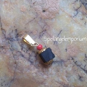 Könnte beinhalten: Ein goldfarbener Anhänger mit einem schwarzen, rechteckigen Stein und einer kleinen roten Perle. Der Anhänger hat eine goldene Öse und liegt auf einer strukturierten, hellbraunen Oberfläche. Der Text "Spellangelemporium" ist sichtbar.