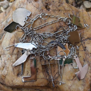 May include: A silver charm bracelet with various charms, including an axe, a boot, and a hook. The charms are connected by a chain. The background is a light brown rock. The text "Spellangelemporium" is in the top right corner.