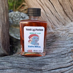May include: A small, rectangular glass bottle filled with a reddish-brown liquid. The label reads "Vandi Parfum" with the words "SHUT UP" and "TAPA BOCA" below a cartoon image. The bottle has a black cap and is set on a weathered wooden surface.