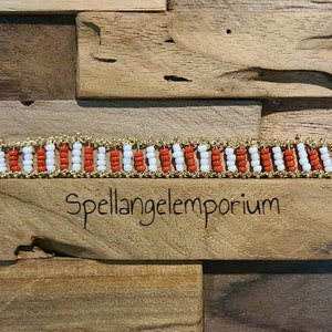 Puede incluir: Una pulsera de cuentas con filas alternas de cuentas rojas y blancas, enmarcada por metal dorado. La pulsera se exhibe sobre una superficie de madera con el texto "Spellangelemporium" grabado en un bloque de madera.