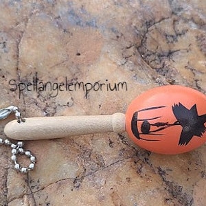 May include: Orange wooden maraca with a black silhouette of a bird in a cage design. The maraca has a wooden handle and a silver chain attached.