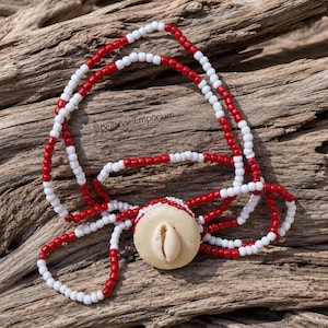 May include: A necklace featuring alternating red and white beads arranged in a multi-strand design. A circular, cream-colored pendant sits at the center, featuring a single white cowrie shell embedded in its surface. The text SpellangelEmporium is visible.